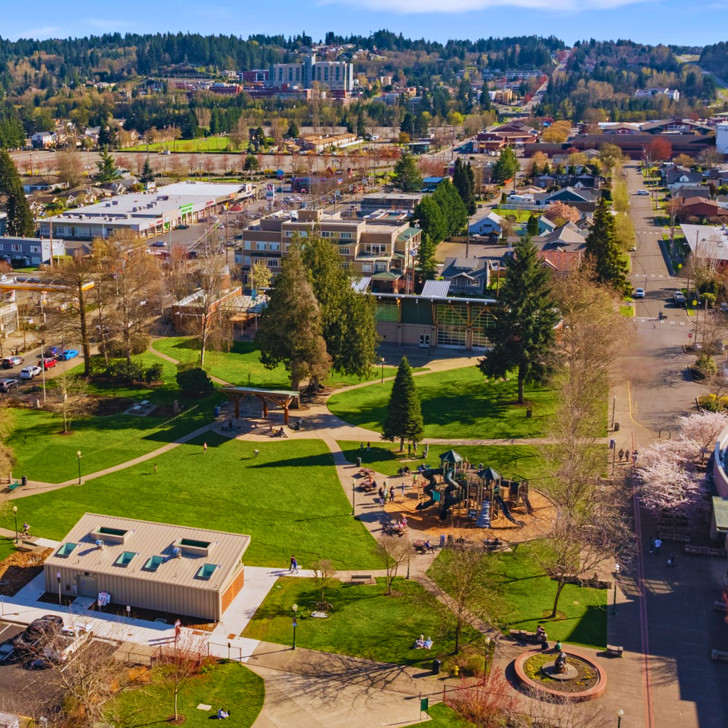 A sunny aerial view of downtown Puyallup, Washington, showcasing a central green park with walking paths, a modern playground, nearby businesses, and neighborhoods—illustrating the community setting where many people search for a dispensary near Puyallup