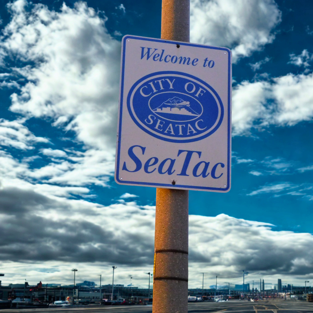 A vertical view of a welcome sign for the City of SeaTac mounted on a light pole, with the airport parking lot and terminal buildings visible in the background under a dramatic, cloud-filled sky—marking the gateway to an area where many travelers search for a dispensary near SeaTac.